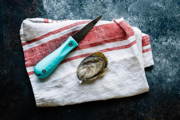 Oyster shucking knife next to oyster on top of tea towel