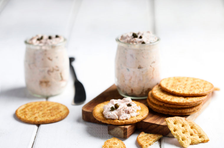 Smoked fish dip in jar next to crackers on top of small cutting board with blurred background
