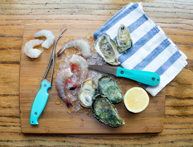 Peeled shrimp, headless shrimp, oysters, lemon, tea towel and shrimp deveiner laying on a wood cutting board