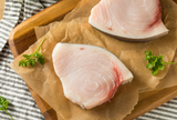 Two fresh swordfish steaks on a wooden cutting board with parsley leaves beside them. thumbnail